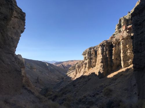 A scenic view of a canyon with rocky cliffs under a clear blue sky, showcasing rugged terrain and distant mountains.