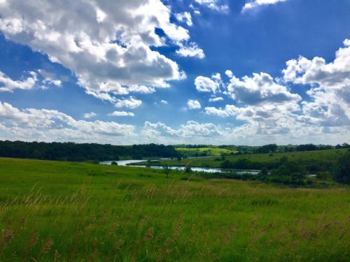 A scenic landscape featuring lush green fields, a river, and a bright blue sky with fluffy white clouds.