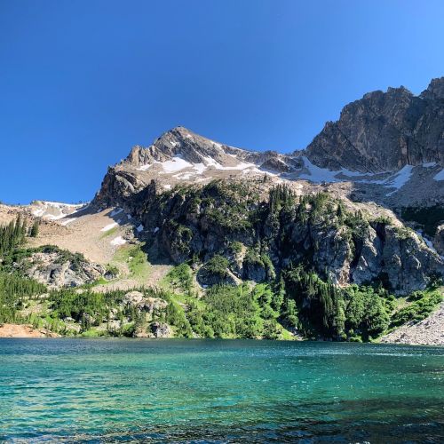 A serene mountain landscape with a clear blue lake, lush greenery, and rocky peaks under a bright blue sky.