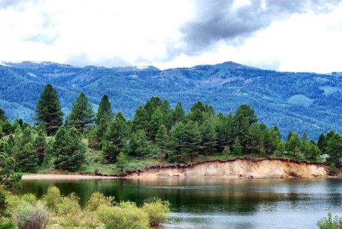 A serene landscape featuring a calm lake surrounded by lush green trees and mountains under a cloudy sky.