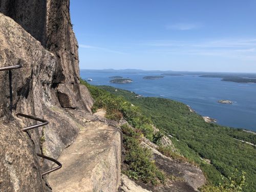 A rocky cliff with metal rungs overlooks a scenic view of a blue bay and green islands under a clear sky.