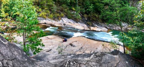 A panoramic view of a rocky riverbank with flowing water, surrounded by lush greenery and trees.