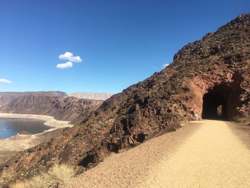 A dirt path leads through a rocky tunnel, with mountains and a lake visible in the background under a clear blue sky.