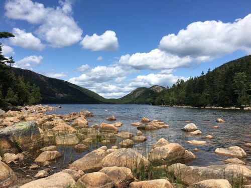 A serene lake surrounded by mountains, with rocky shores and fluffy clouds in a blue sky.