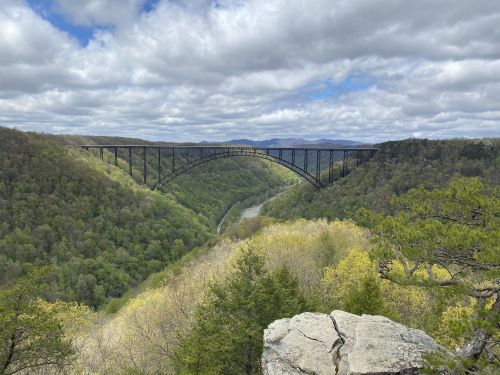 A large metal bridge spans a lush green valley, with rolling hills and a river below, under a cloudy sky.