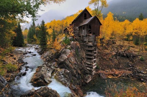 A rustic wooden cabin perched on a rocky outcrop beside a flowing river, surrounded by vibrant autumn foliage.