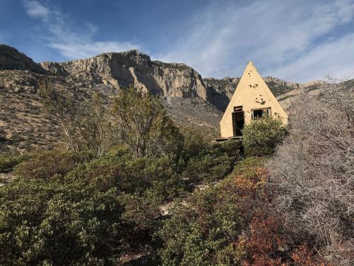 An abandoned triangular house surrounded by shrubs and mountains under a blue sky.