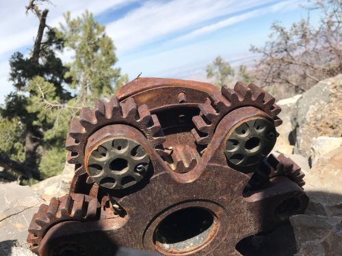 Rusty mechanical gears resting on rocky terrain with a scenic view of trees and sky in the background.