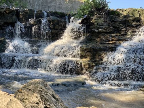 A cascading waterfall flows over rocky terrain, surrounded by greenery under a clear blue sky.