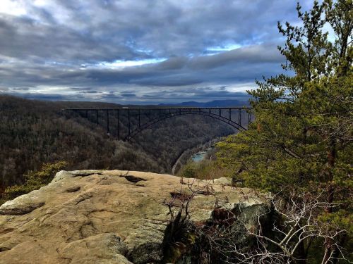 A scenic view of a bridge spanning a valley, surrounded by trees and mountains under a cloudy sky.