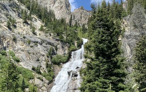 A cascading waterfall flows down rocky cliffs, surrounded by lush green trees and mountains under a blue sky.