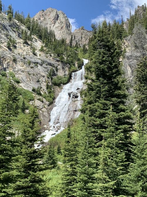 A cascading waterfall flows down rocky cliffs, surrounded by lush green trees and mountains under a blue sky.