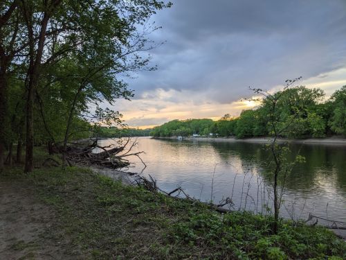 A serene river scene with trees lining the bank, reflecting clouds and a sunset in the water.
