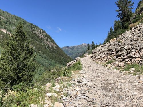 A rocky trail winds through a lush green valley, surrounded by mountains under a clear blue sky.