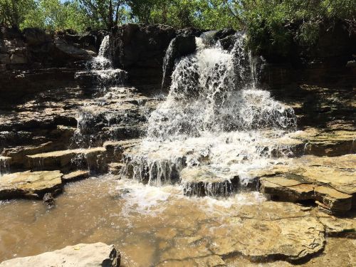 A cascading waterfall flows over rocky terrain, surrounded by lush greenery and clear water pools.