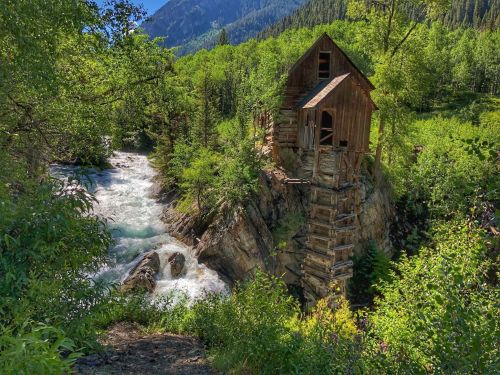 A rustic wooden cabin perched on a rocky cliff beside a rushing river, surrounded by lush green trees and mountains.
