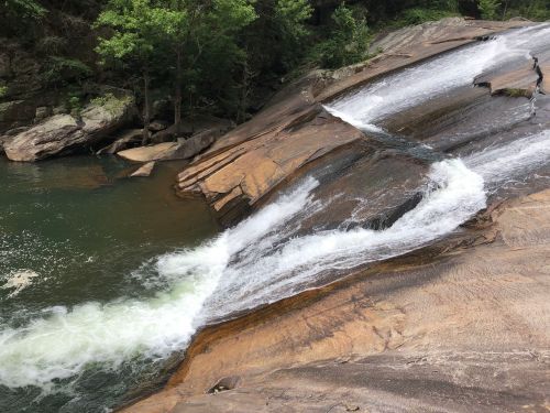 A cascading waterfall flows over smooth, rocky terrain into a serene green pool surrounded by trees.