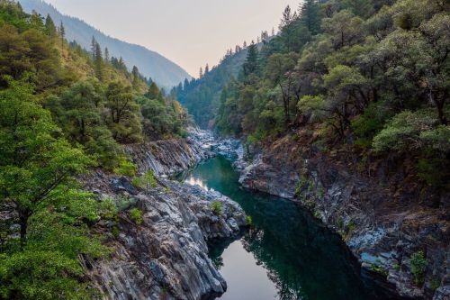 A serene river flows through a rocky canyon, surrounded by lush green trees and mountains in the background.