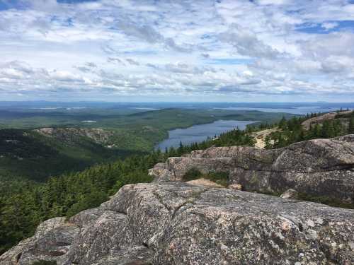 A scenic view from a rocky summit overlooking a lake surrounded by lush green forests and a cloudy sky.