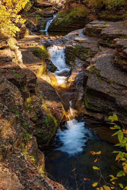 A serene waterfall cascades over rocky terrain, surrounded by autumn foliage and tranquil pools of water.