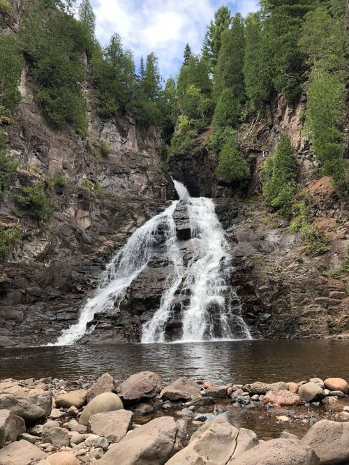 A cascading waterfall flows down rocky cliffs, surrounded by lush green trees and a serene pool at the base.