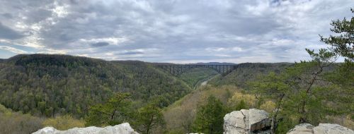 A panoramic view of a bridge spanning a lush green valley, surrounded by rolling hills and a cloudy sky.