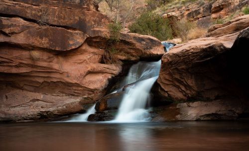 A serene waterfall cascades over rocky cliffs into a calm pool, surrounded by earthy tones and lush vegetation.