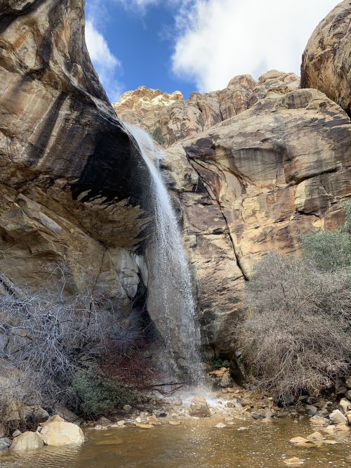 A waterfall cascading down rocky cliffs, surrounded by greenery and a clear blue sky with scattered clouds.