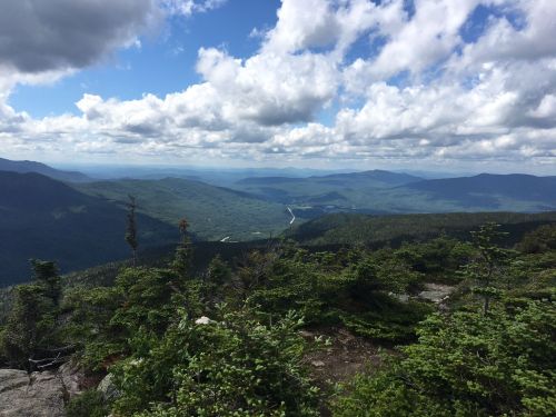 A panoramic view of rolling mountains and valleys under a partly cloudy sky, with lush greenery in the foreground.