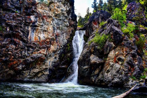 A waterfall cascades down rocky cliffs into a serene pool, surrounded by lush greenery and rugged terrain.