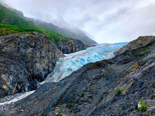 A glacier flows down rocky terrain, surrounded by green hills and a cloudy sky.