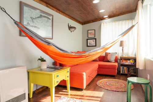 A cozy living room featuring an orange hammock, a red sofa, colorful decor, and natural light from a window.