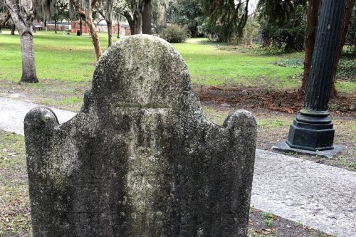 A weathered gravestone in a grassy park, surrounded by trees and a lamppost in the background.