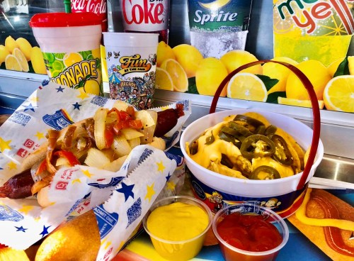 A colorful food display featuring a hot dog with onions, nachos with jalapeños, and various drinks in the background.