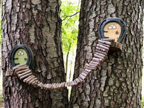 Two tree trunks with whimsical doors and a bridge made of sticks connecting them. Lush greenery in the background.