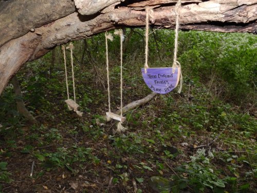 A wooden sign reading "There Carefully Fairies Play Here" hangs from a tree branch above small swings in a forested area.
