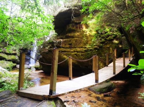 A wooden bridge with chains crosses a serene stream in a lush, green forest with rocky formations and a waterfall.