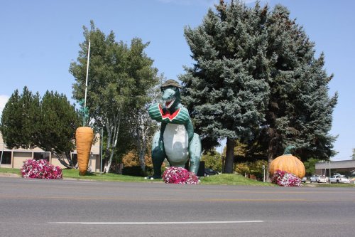A large dinosaur statue wearing a hat stands among oversized vegetables and colorful flowers by the roadside.