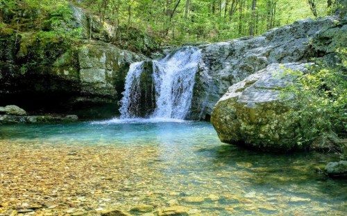 A serene waterfall cascades into a clear pool surrounded by lush greenery and rocky terrain.