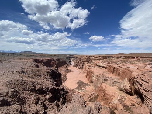 A vast canyon landscape with rocky cliffs, a river below, and a blue sky dotted with fluffy clouds.