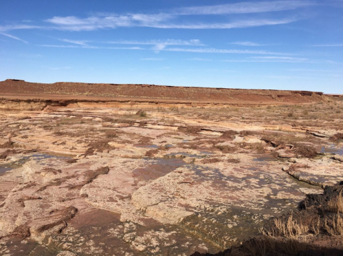 A dry, rocky landscape with layered earth formations under a clear blue sky. Sparse vegetation is visible.