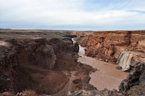 A wide canyon with steep, rocky cliffs and a winding river flowing through the landscape under a cloudy sky.