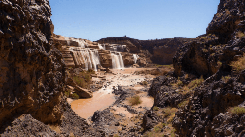 A scenic view of a waterfall cascading over rocky cliffs into a sandy riverbed, surrounded by rugged terrain.