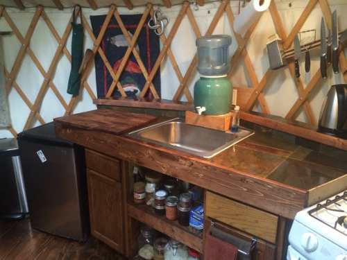 A cozy kitchen in a yurt featuring a sink, wooden countertops, a water dispenser, and organized shelves with jars.
