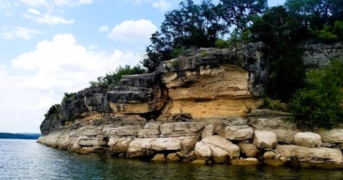 Rocky shoreline with layered cliffs and greenery under a partly cloudy sky, reflecting in calm water.