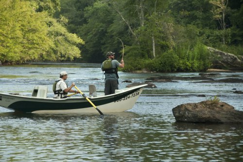 Two men in a small boat fishing on a calm river surrounded by lush greenery and rocks.