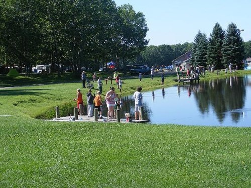 Children and adults fishing from a dock by a calm pond, surrounded by trees and grassy areas on a sunny day.
