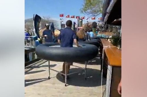 A person carries a large inflatable tube on a dock, with flags and people in the background.