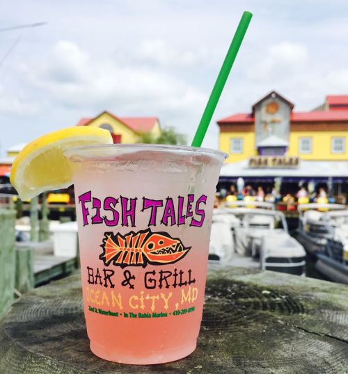 A refreshing drink with a lemon slice in a cup from Fish Tales Bar & Grill, with boats and a colorful building in the background.