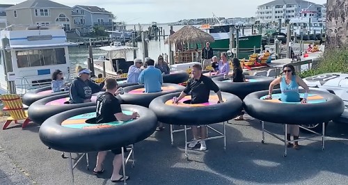 A group of people enjoying a fun outdoor activity on inflatable trampolines by a marina with boats in the background.